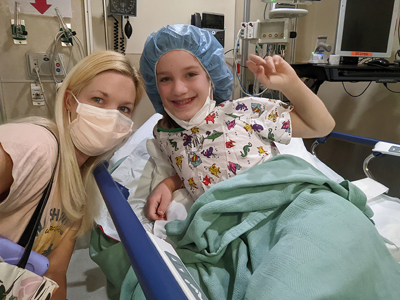 Una mujer rubia con una mascarilla se agacha junto a una niña sonriente acostada en una cama de hospital.