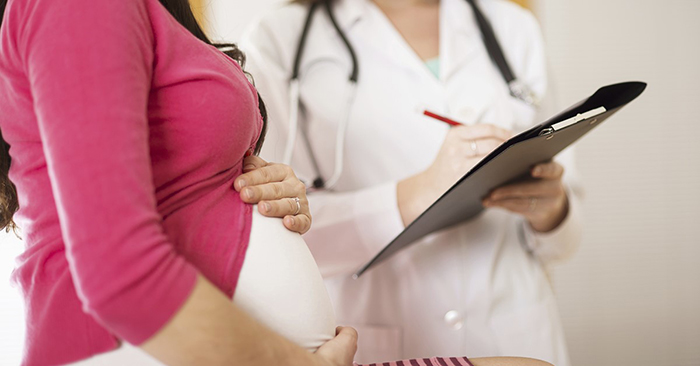 Imagen recortada de una mujer embarazada y su proveedor de atención médica, con la imagen enfocada en el vientre de la mujer y el portapapeles del proveedor.