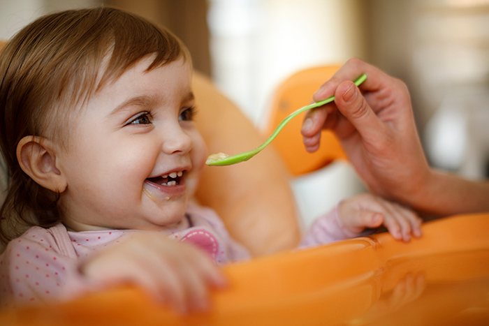 Bebé en silla alta que sonríe y a la que le dan de comer alimentos para bebé en una cuchara.