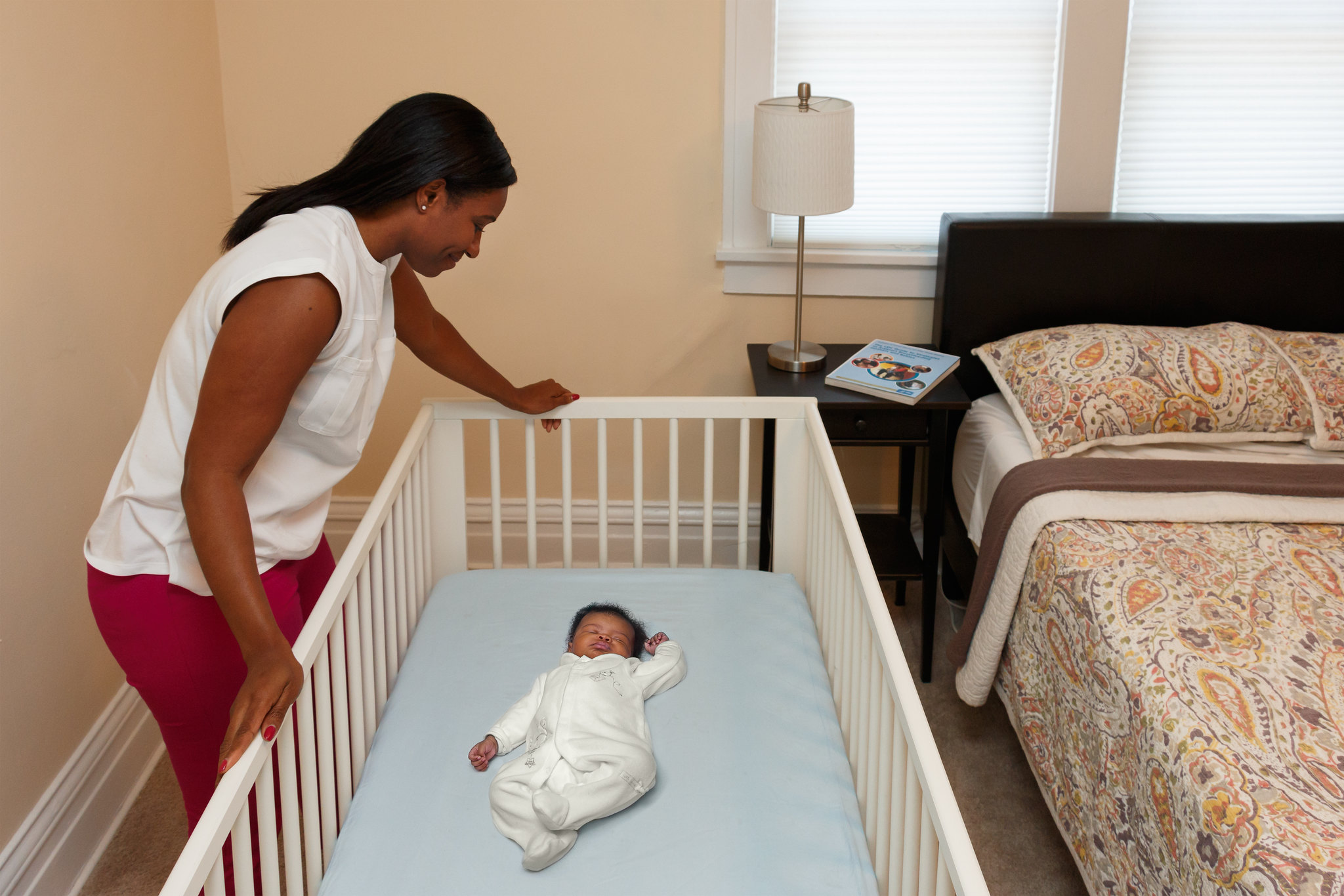 Una mujer mira a un bebé que duerme boca arriba en una cuna que no incluye ropa de cama blanda o suelta, protectores de cuna, juguetes u otros objetos en el área para dormir.