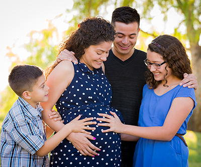 Familia reunida alrededor de una mujer embarazada. La hija y el hijo de la mujer embarazada se sujetan de las manos alrededor de su estómago.