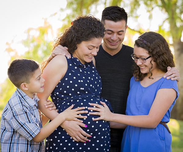 Familia reunida alrededor de una mujer embarazada. La hija y el hijo de la mujer embarazada se sujetan de las manos alrededor de su estómago.