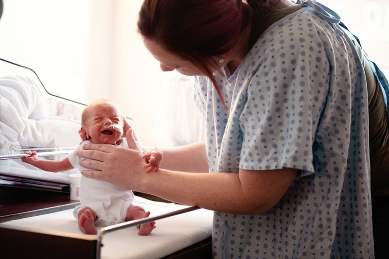 Mujer en bata de hospital sostiene a un bebé prematuro sentado en la mesa.