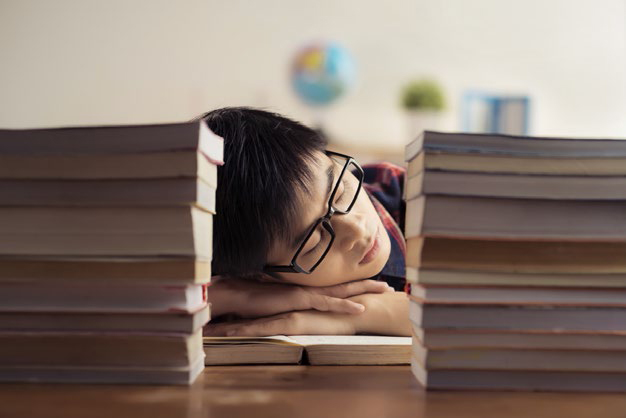 Varón adolescente descansando su cabeza sobre un escritorio entre dos pilas de libros.