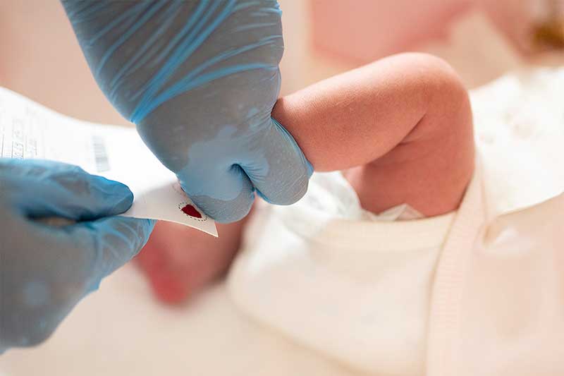 Unas manos enguantadas sostienen el talón de un bebé, con una mancha de sangre visible en el papel de filtro.