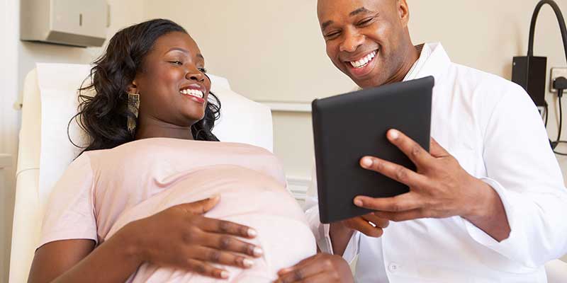 Una mujer embarazada y su proveedor de atención médica sonríen ante una tableta, que presumiblemente contiene resultados de pruebas u otra información de salud.