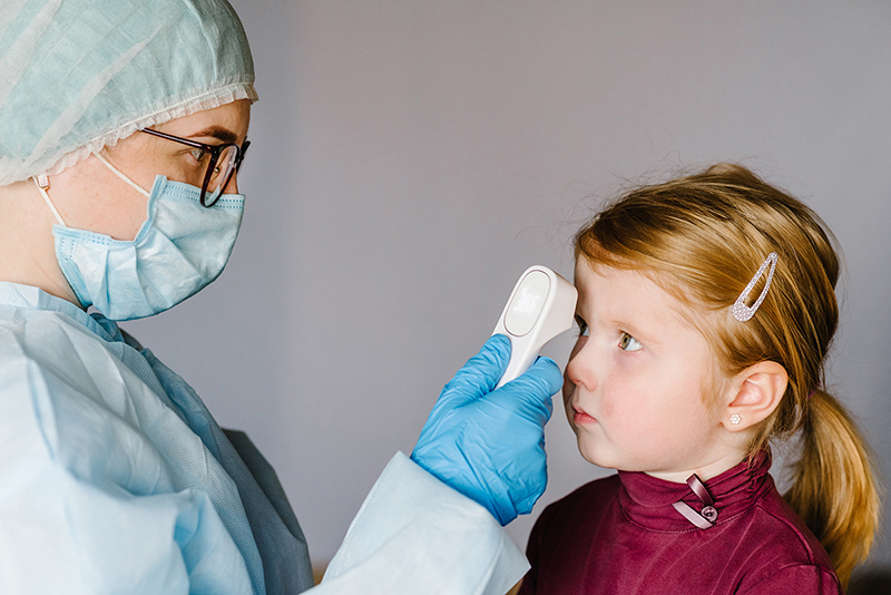Un profesional médico con máscara y guantes examina la frente de un niño con un termómetro digital.