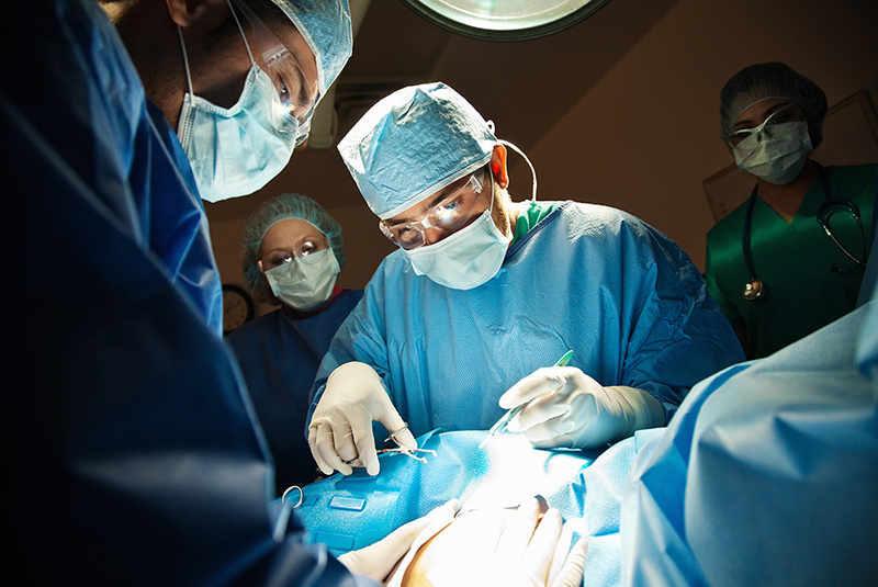 Médicos y enfermeras con vestimenta quirúrgica de pie ante el torso cubierto de un paciente. 