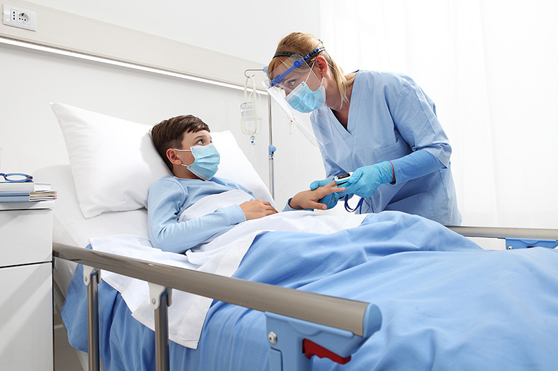 Niño con mascarilla acostado en la cama de un hospital mientras la enfermera controla el nivel de oxígeno en un oxímetro de pulso de dedo.