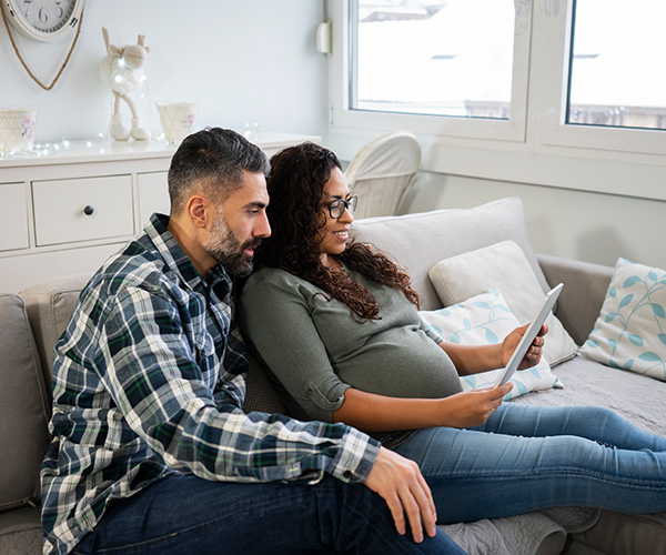 Un hombre y una mujer embarazada sentados juntos en un sofá miran algo en la tableta que sostiene la mujer.