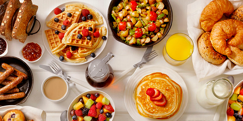 Una mesa blanca con una variedad de bebidas y platos de alimentos para el desayuno, incluidos waffles, panqueques, frutas y croissants.