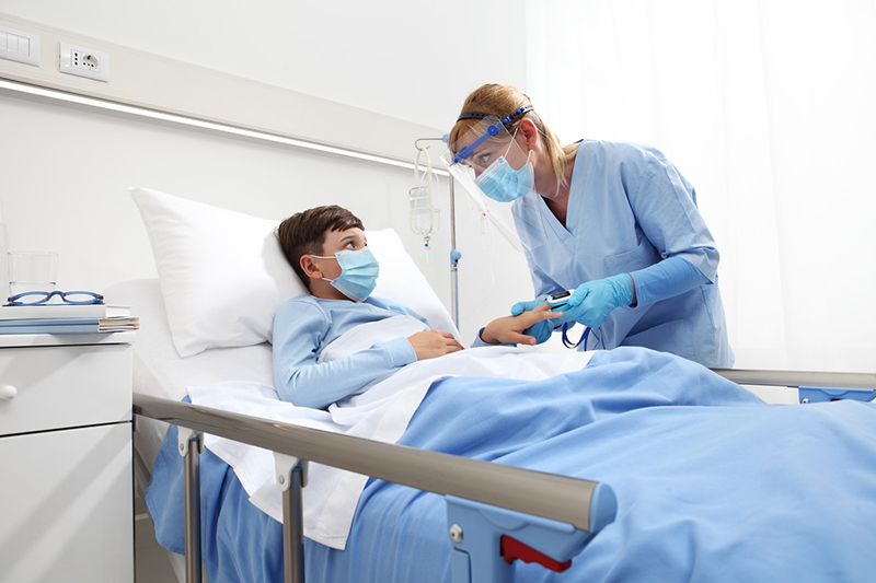 Niño con mascarilla acostado en la cama de un hospital mientras el proveedor de atención médica controla el nivel de oxígeno en un oxímetro de pulso de dedo.