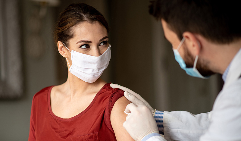 Una mujer recibe una vacuna en la parte superior del brazo. Ella y la persona que administra la inyección están enmascarados.