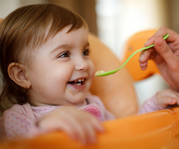 Una bebé sonríe en su sillita alta mientras un adulto, que no se ve en la imagen, la alimenta con una cuchara verde.