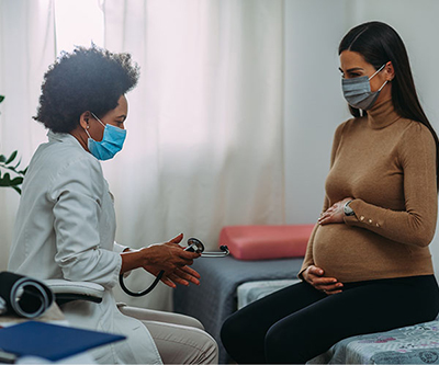 Una embarazada abraza su vientre mientras habla con su proveedora de atención médica, que sostiene un estetoscopio. Las dos llevan mascarilla.