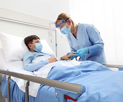 Niño con mascarilla en una cama de hospital que mira a la proveedora de atención médica mientras le mide los signos vitales.  La proveedora tiene mascarilla y un protector facial.