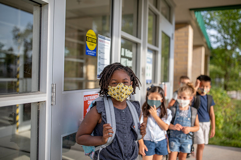 Un grupo de niños en edad escolar hacen una fila frente a un edificio. Todos llevan mascarillas de tela.