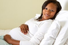 Una mujer de raza negra embarazada sonríe mientras se reclina en la cama con las manos sobre el vientre.