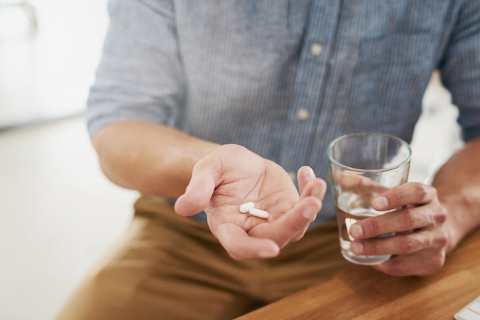 Un hombre sentado en una mesa sostiene pastillas en una mano abierta mientras sostiene un vaso de agua en la otra mano.