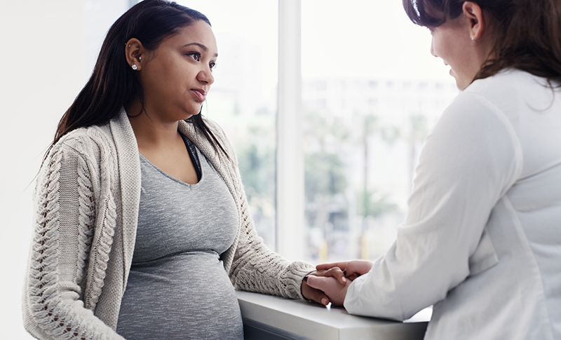 Un proveedor de atención médica que lleva una bata blanca sostiene la mano de una persona embarazada mientras habla con ella.