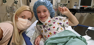 Una mujer rubia con una mascarilla se agacha junto a una niña sonriente acostada en una cama de hospital.