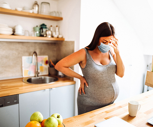Una mujer embarazada usa una mascarilla y parece visiblemente cansada en la cocina.