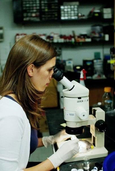 Una mujer de cabello castaño con una chaqueta blanca y guantes blancos de laboratorio mira por el microscopio. Los frascos y otros equipos de investigación científica se ven borrosos en el fondo.