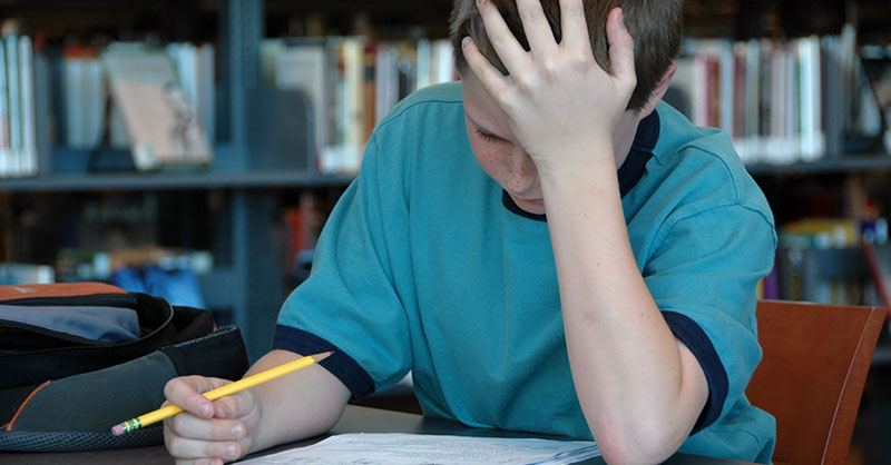 Un adolescente está sentado en una mesa frente a una estantería. Está encorvado sobre un papel. Sostiene un lápiz en la mano derecha y tiene la mano izquierda apoyada en la frente. 