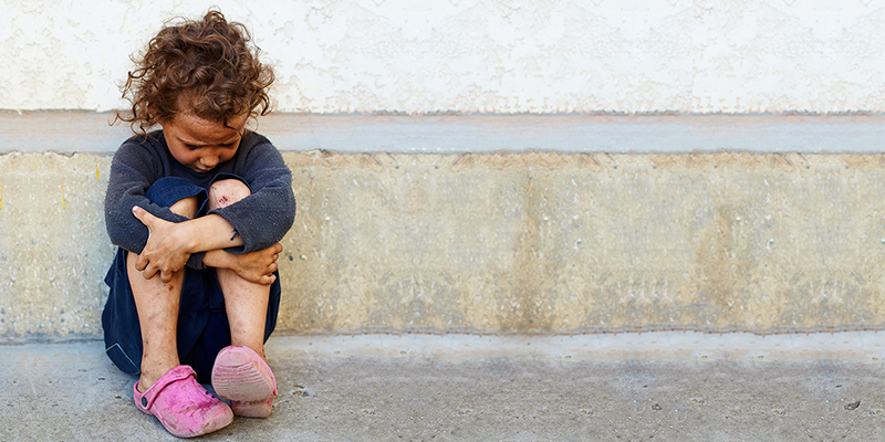 Una niña sentada contra un edificio, sosteniendo sus rodillas y mirando hacia abajo.