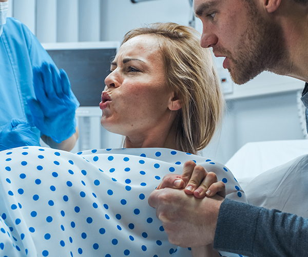 Una mujer embarazada que se encuentra en trabajo de parto respira con dificultad, mientras su marido le sostiene la mano y un profesional sanitario con mascarilla la observa.