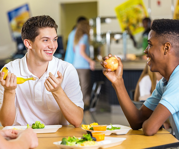 Dos adolescentes sonrientes. Uno sostiene un plátano y el otro una manzana; están sentados en una mesa llena de platos de verduras. 