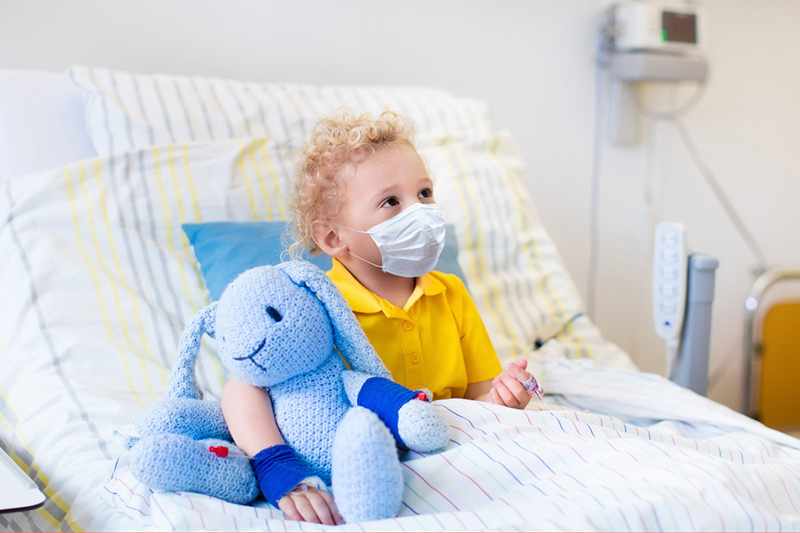 Un niño se sienta en una cama de hospital. Lleva una mascarilla y sostiene un conejito de peluche azul. 