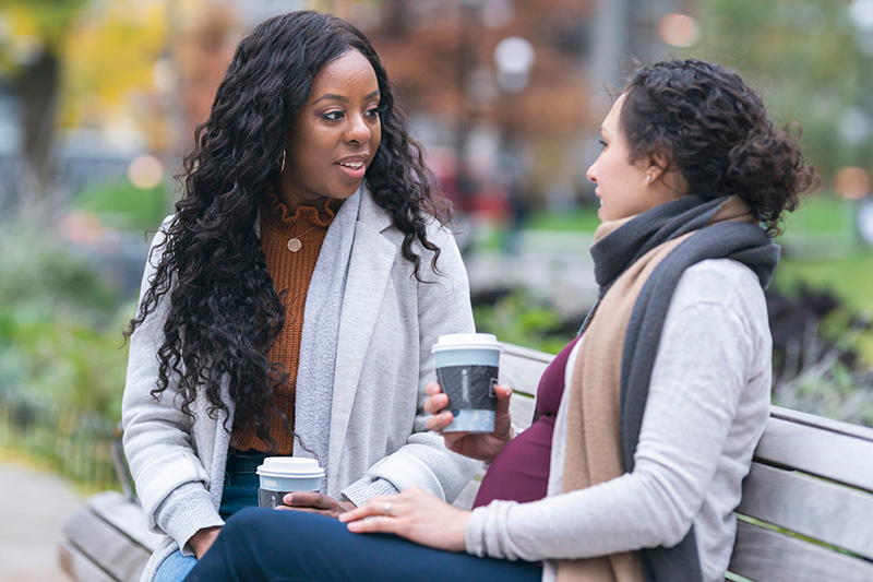 Una mujer negra y su amiga blanca embarazada se sientan en un banco del parque tomando café y hablando.