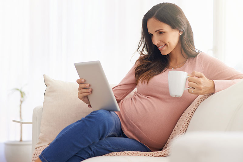 Una mujer embarazada se sienta en un sofá leyendo una tableta y sosteniendo una taza en la mano.