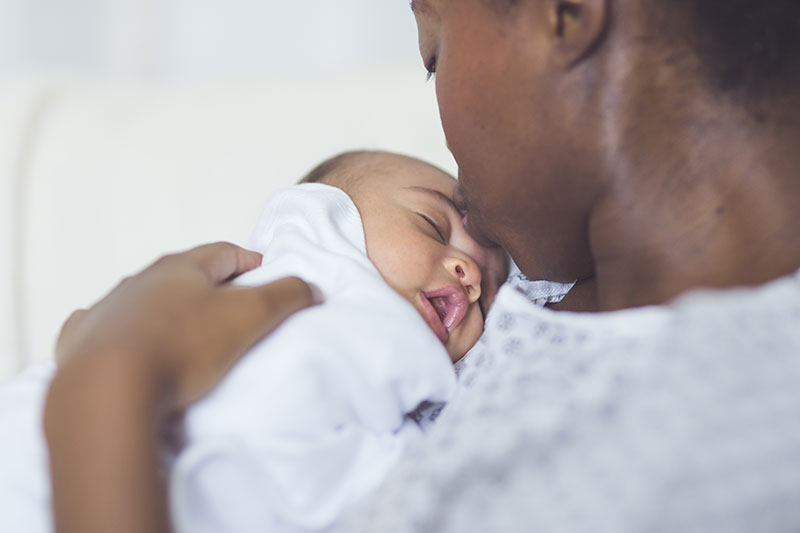 Una mujer negra con bata de hospital besa la frente de su bebé recién nacido.