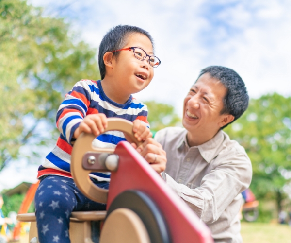 Un padre y su hijo, que tiene síndrome de Down y usa anteojos, juegan juntos en un parque infantil al aire libre. Ambos sonríen.