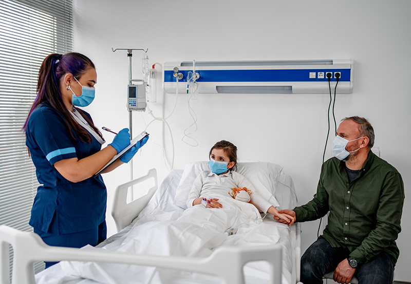 Niño en una cama de hospital, trabajador de la salud a la izquierda escribiendo en un portapapeles, padre sentado a la derecha. Todos utilizan mascarillas.