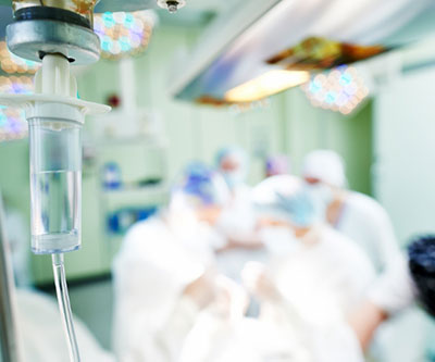 Una cámara de goteo intravenoso aparece nítidamente en primer plano, con el personal médico y el paciente borrosos en el fondo