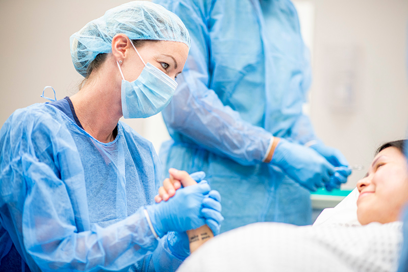 Trabajador de la salud con ropa quirúrgica sostiene la mano de una mujer embarazada acostada en una camilla. En el fondo, se ve el torso superior de un trabajador de la salud con ropa quirúrgica.