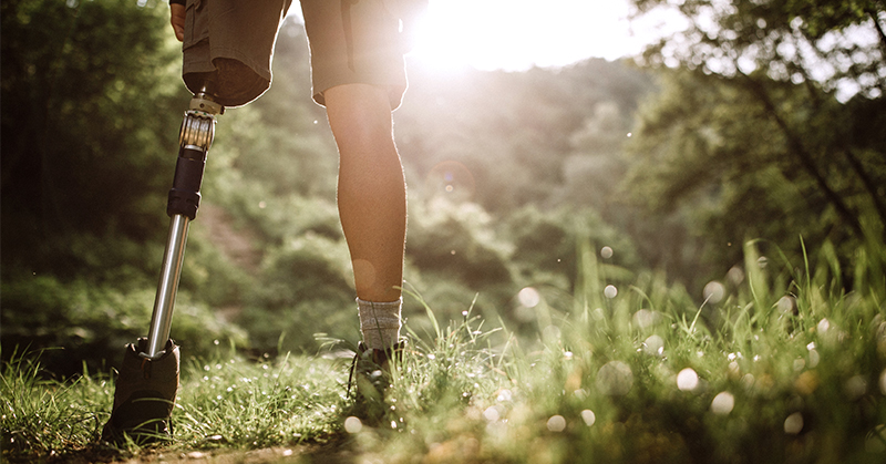 Una persona con una prótesis de parte inferior de la pierna se encuentra en un sendero cubierto de hierba. En el fondo, se ven los árboles y el sol.