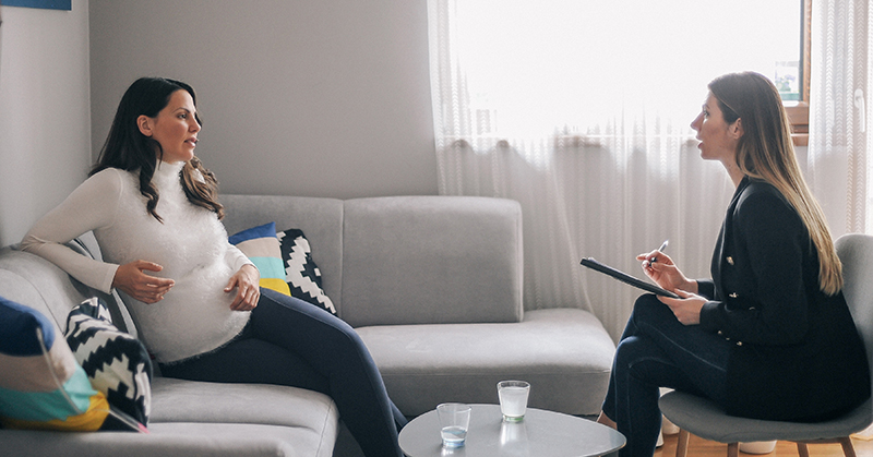 Persona embarazada, sentada en el sofá, hablando con un profesional de la salud mental con un bloc de notas y un bolígrafo.
