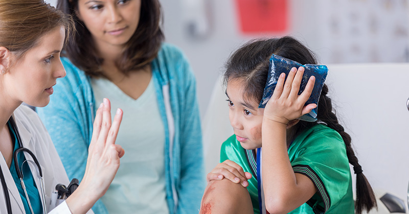 Un trabajador de la salud levanta tres dedos frente a un niño que apoya una bolsa de hielo contra su cabeza mientras sus padres observan.