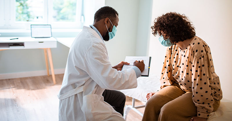 Un proveedor de atención médica con mascarilla y bata blanca de laboratorio escribe en un portapapeles mientras un paciente con mascarilla sentado en una mesa de examinación observa.