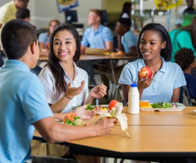 Un grupo diverso de adolescentes hombres y mujeres almorzando en la cafetería de una escuela. En las mesas, delante de ellos, hay platos llenos de verduras y frutas.