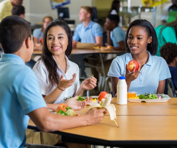 Un grupo diverso de adolescentes hombres y mujeres almorzando en la cafetería de una escuela. En las mesas, delante de ellos, hay platos llenos de verduras y frutas.