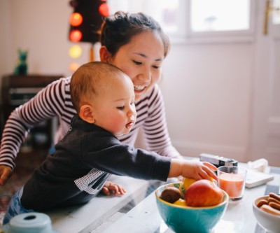 Una joven madre asiática con una camiseta a rayas en una encimera con su bebé. El bebé intenta alcanzar una fruta que hay en un recipiente sobre la encimera.