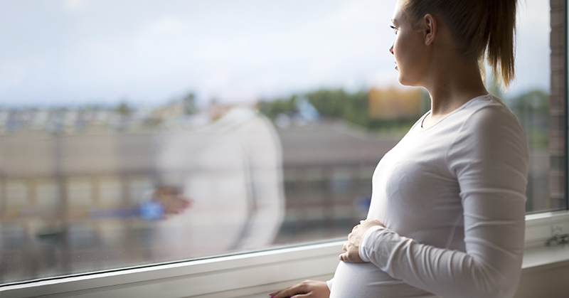 Una persona embarazada mira por la ventana con una mano en el vientre.