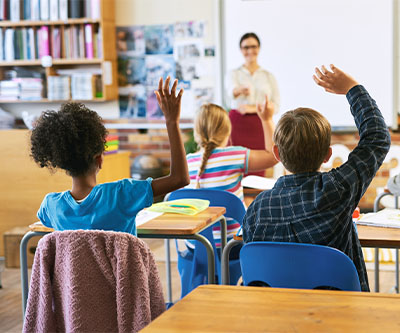 Una vista posterior de un grupo de niños en un salón de clases que tienen las manos levantadas mientras miran a un maestro con gafas, que está parado al frente de la clase.