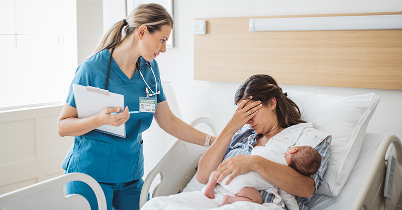 Un médico al lado de la cama de una persona que recientemente ha dado a luz con una mano sobre los ojos, sosteniendo al recién nacido.