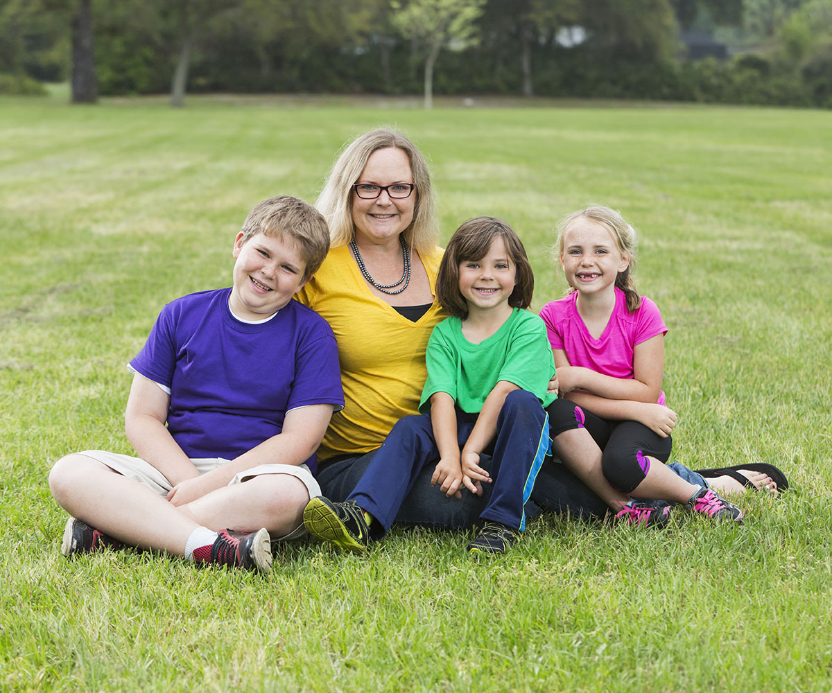 Un adulto y tres niños sentados juntos en un campo cubierto de hierba, sonriendo y mirando al frente en posición de pose.